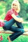 Olivia Newton-John wearing jeans and a red shirt and sitting on a wooden fence