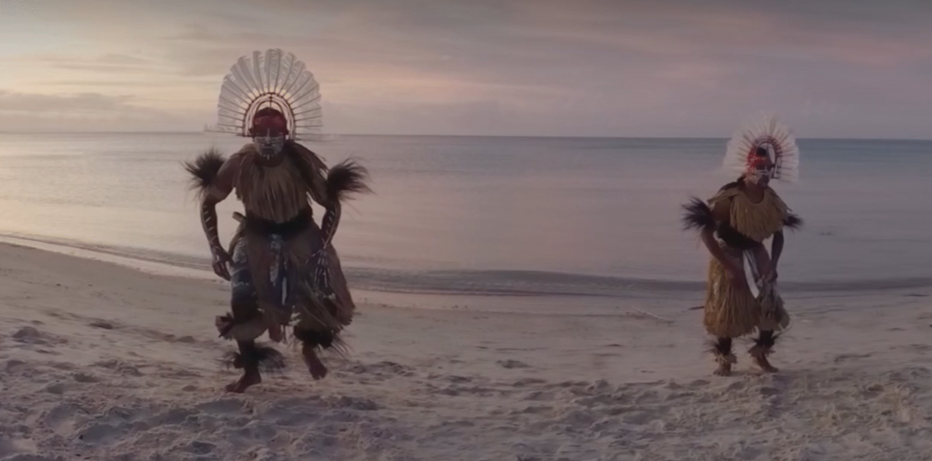 Two Indigenous men in traditional costume dancing on a beach.