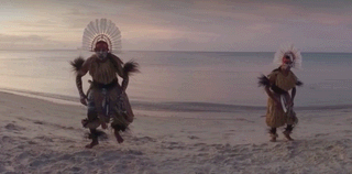 Two Indigenous men in traditional costume dancing on a beach.