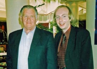 Two men dressed in shirts and suit jackets pictured from the waist up, smiling at camera.