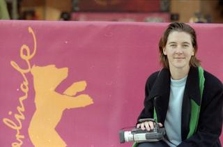 A young woman in front of a pink banner holding a hand-held video camera.