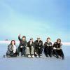 Six people sit outside on a flat surface. They are laughing and smiling and one of them is raising his hand to the sky.