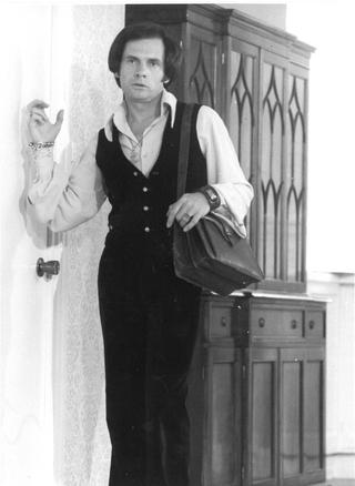 Person in a buttoned vest over a collared shirt, carrying a shoulder bag, standing near a door with a cabinet behind.