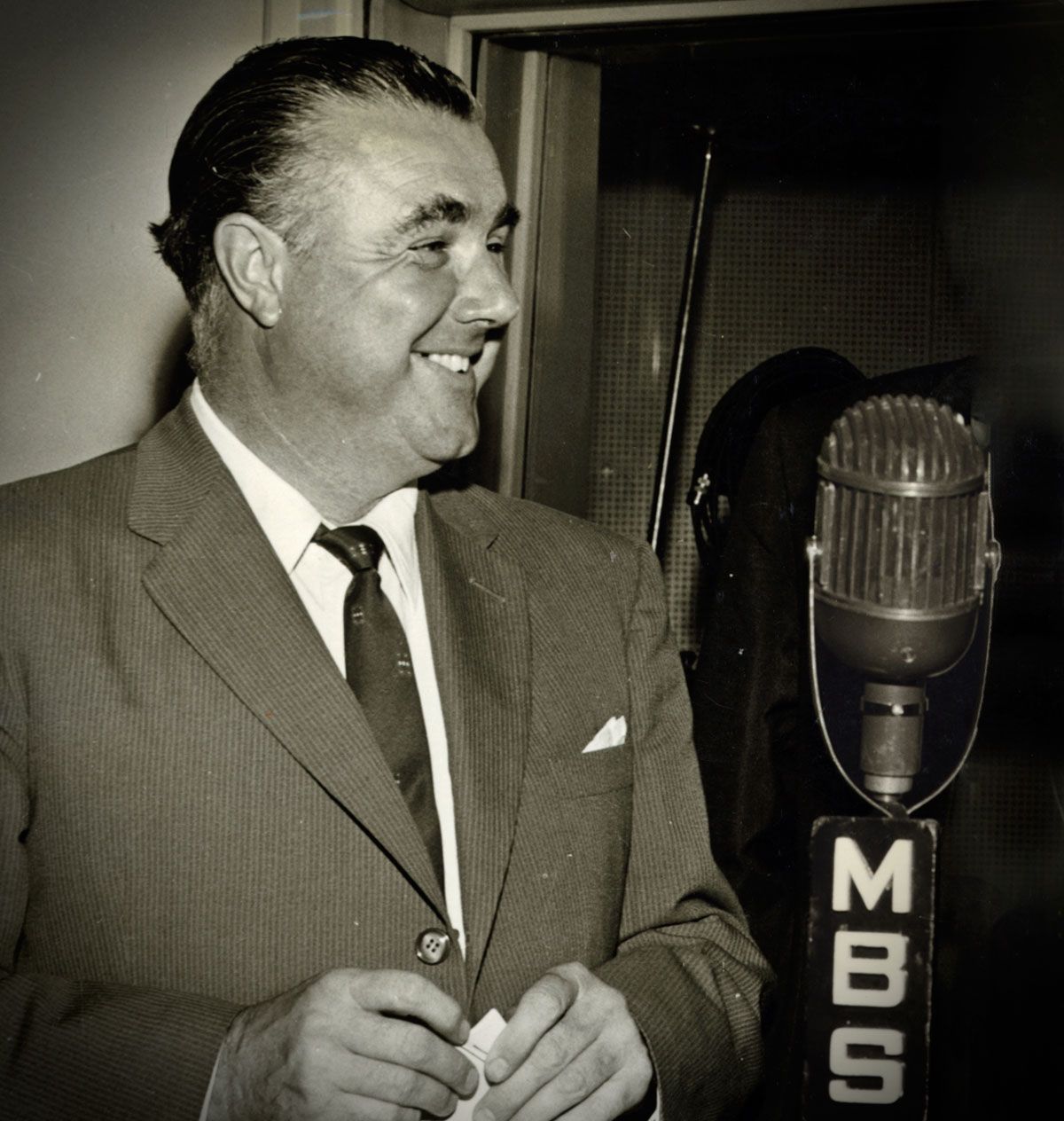 Radio announcer Terry Dear, circa 1960, standing next to a microphone with the MBS logo on it. He is looking sideways and smiling.