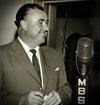 Radio announcer Terry Dear, circa 1960, standing next to a microphone with the MBS logo on it. He is looking sideways and smiling.