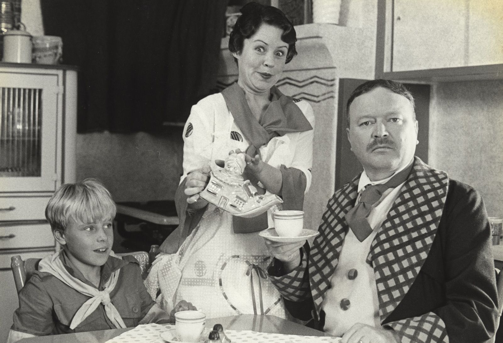 A young boy and his parents sitting around a table, dressed in costumes from c 1930s in a scene from the film Fatty Finn