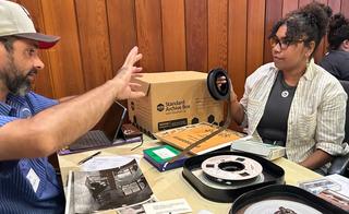 A man and a woman sitting opposite each other at a desk with film reels and paperwork in front of them.