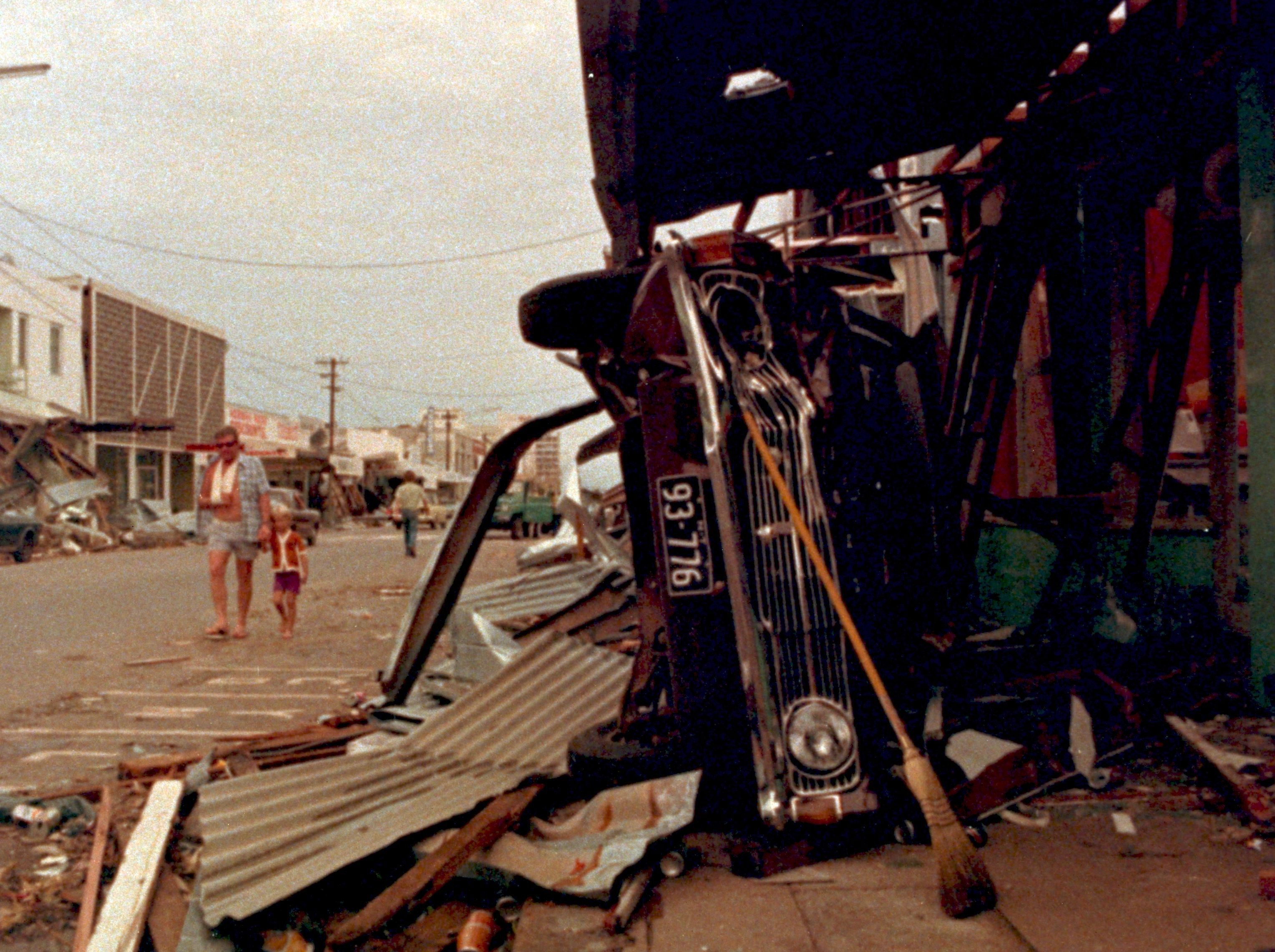 A street in Darwin that has been destroyed by Cyclone Tracy in 1974. You can see debris all over the road and a car on its side.
