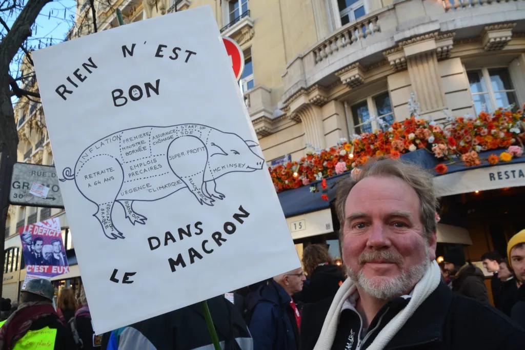 En eldre mann med hvitt hår og skjegg som smiler. I hånden holde han en plakat med teksten "Rien n'est bon dans le Macron" me bilde av en gris. I bakgrunnen ser man mennesker som går i demonstrasjonstog i en by. 