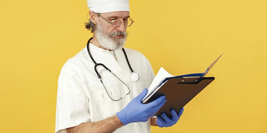 A Medicare doctor reviews patient medical records on a clipboard, wearing white scrubs, a surgical cap, and blue gloves.