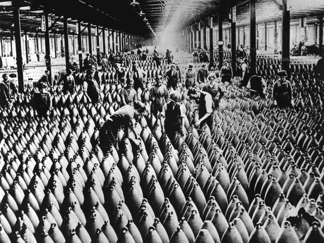 A historic black and white photograph of female munitions workers in headscarves and uniforms standing in front of a wooden factory building in WWI.