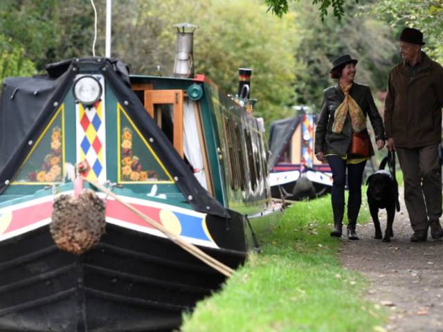 A person walking along the grassy towpath of the Oxford Canal in Banbury with traditional narrowboats moored on the calm water under a bright sky.
