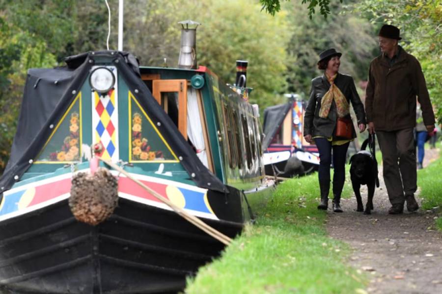 A person walking along the grassy towpath of the Oxford Canal in Banbury with traditional narrowboats moored on the calm water under a bright sky.