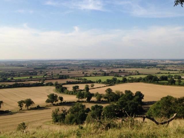 A beautiful panoramic view of the North Oxfordshire countryside featuring rolling green hills and historic stone buildings under a bright and clear sky.
