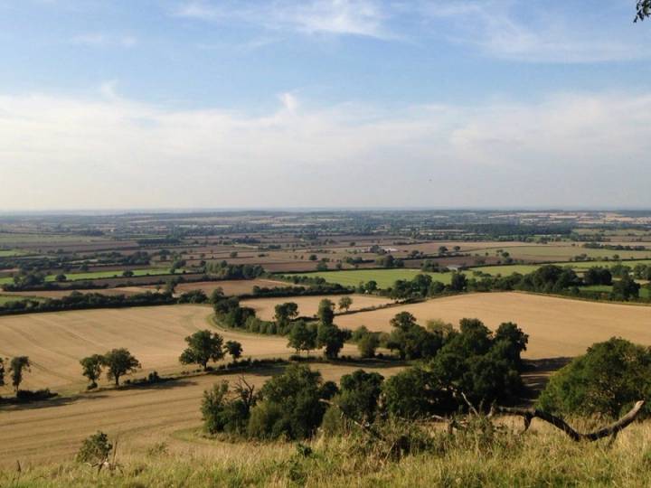 A beautiful panoramic view of the North Oxfordshire countryside featuring rolling green hills and historic stone buildings under a bright and clear sky.