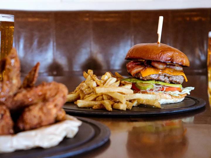 A close up shot of a gourmet beef burger with melted cheese and fresh lettuce in a brioche bun served with a side of golden skin-on chips on a dark plate.