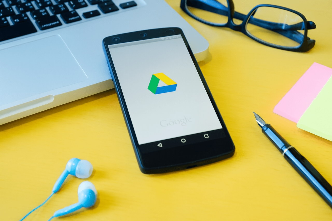 Smartfon z logo Google Drive obok laptopa, okularów i słuchawek na żółtym tle