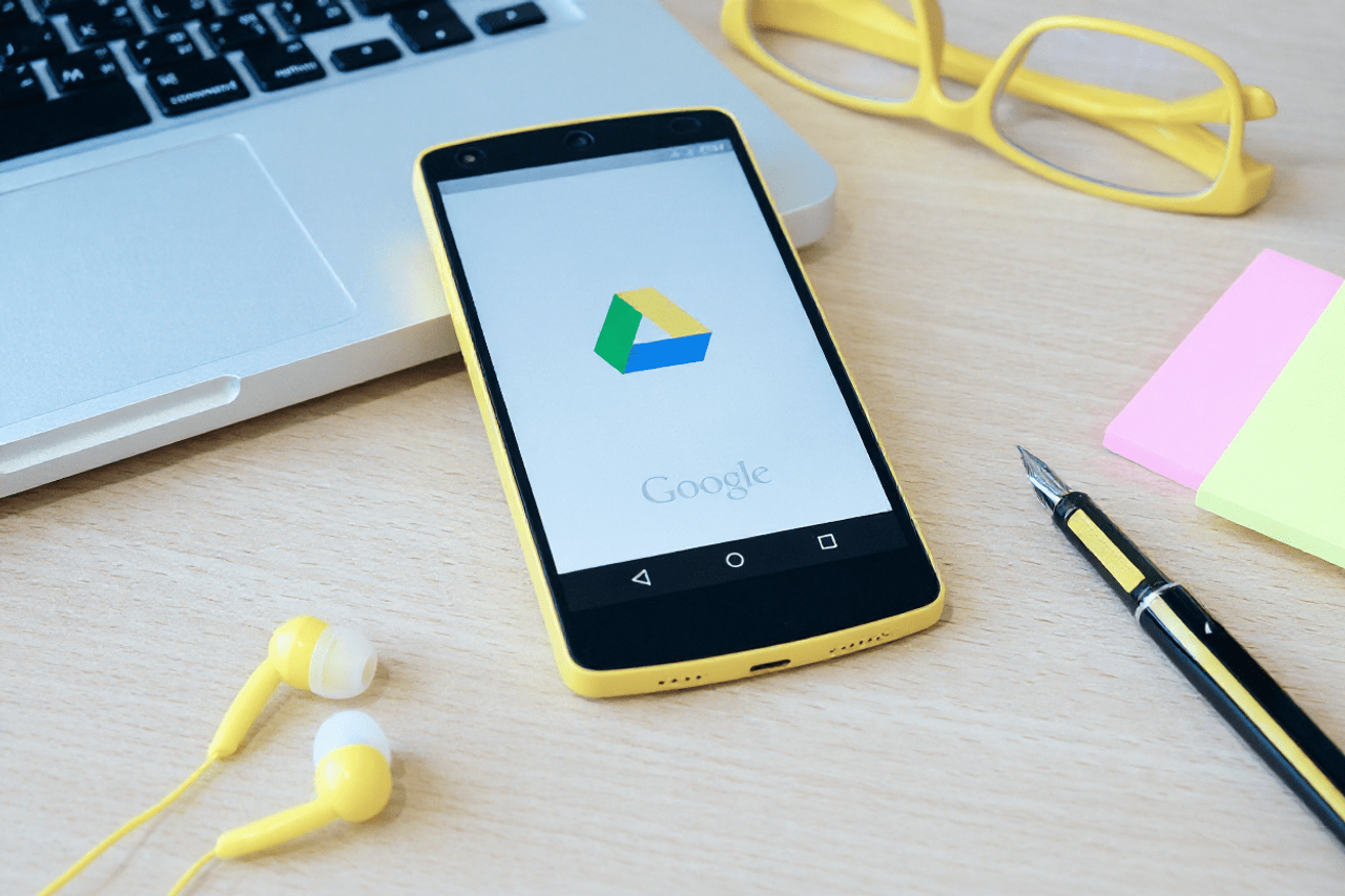 Smartfon z logo Google Drive obok laptopa, okularów i słuchawek na biurku