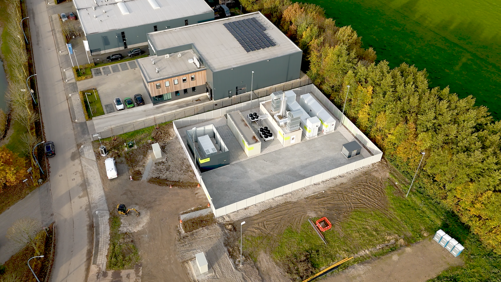 Drone foto van een bedrijventerrein. Rechts is het heel groen, met water en een bos vol bomen. In het midden staan het bedrijventerrein, met gras en bruine aarde eromheen. Links van het bedrijventerrein is een autoweg te zien en een parkeerplaats met twee witte busjes.