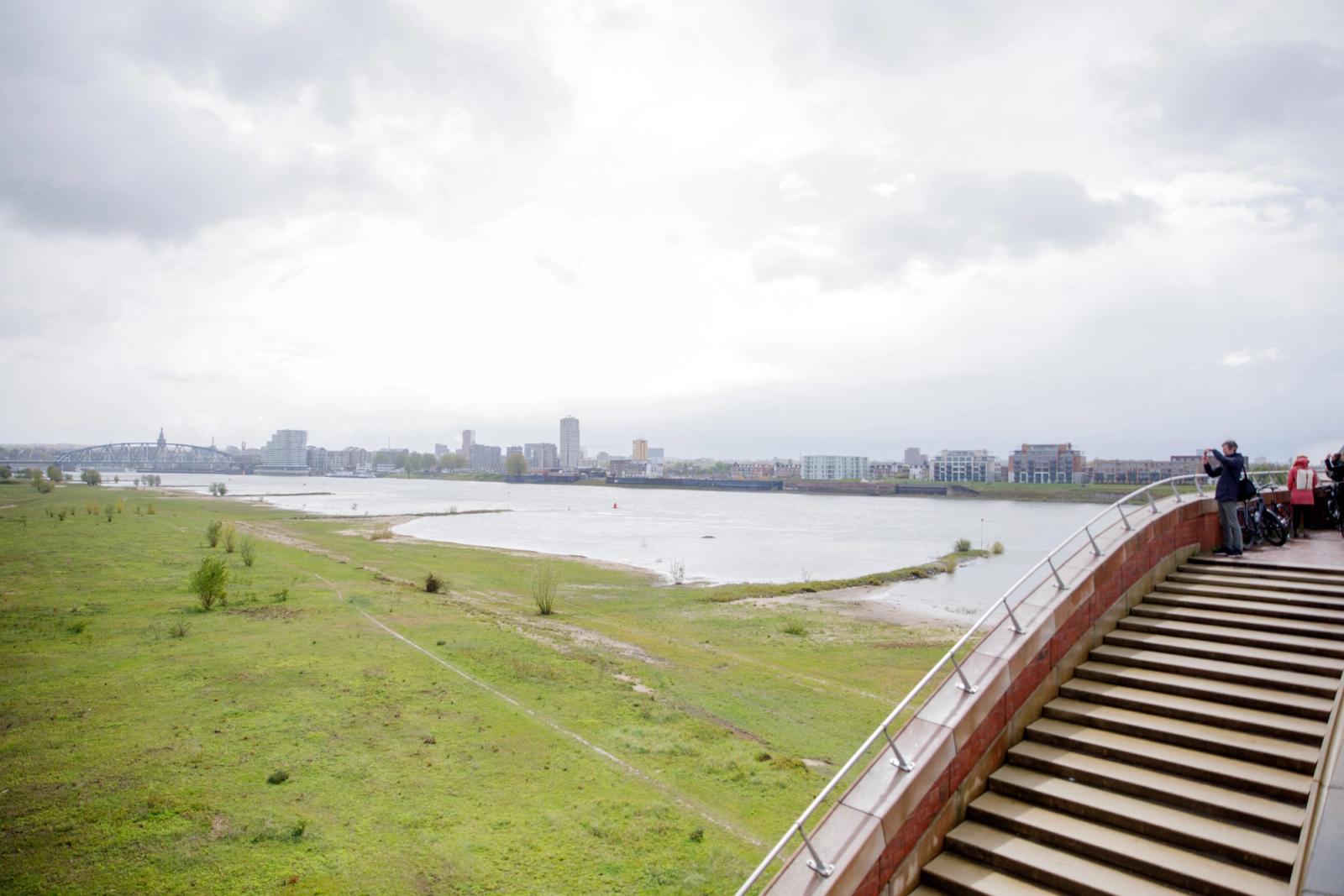 Een uitzicht vanaf een brug bij Rivierpark Nijmegen. Links een groot grasveld, in het midden rivier de Waal en rechts de brug. 
