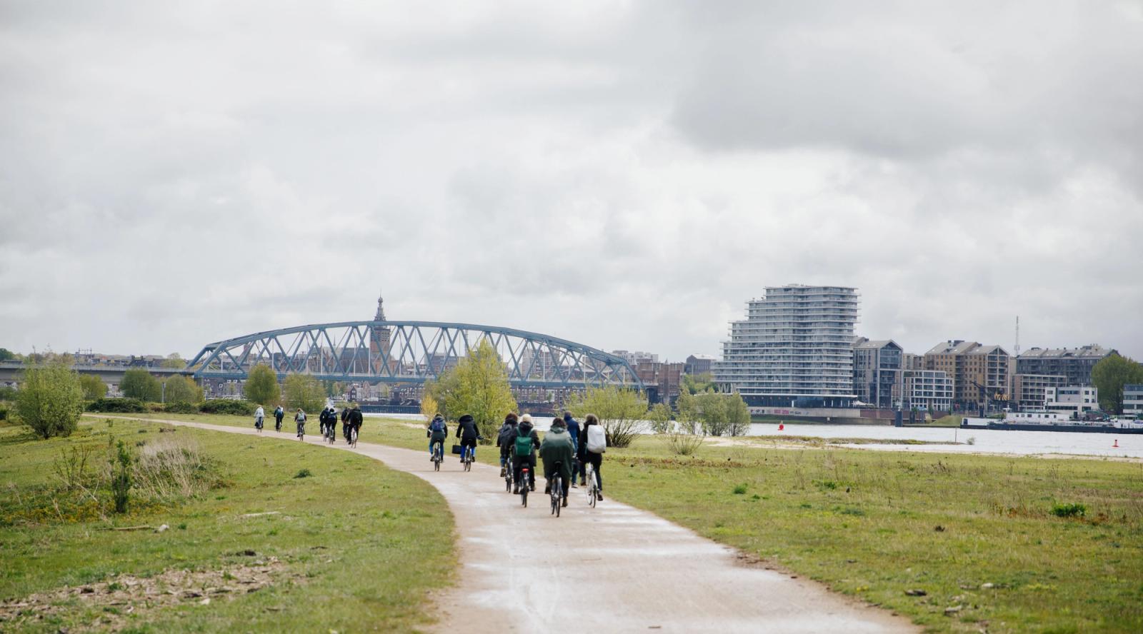 Een fietspad loopt tussen twee grasvelden door. Op het fietspad fietst een groepje fietsers. Op de achtergrond staat een brug.