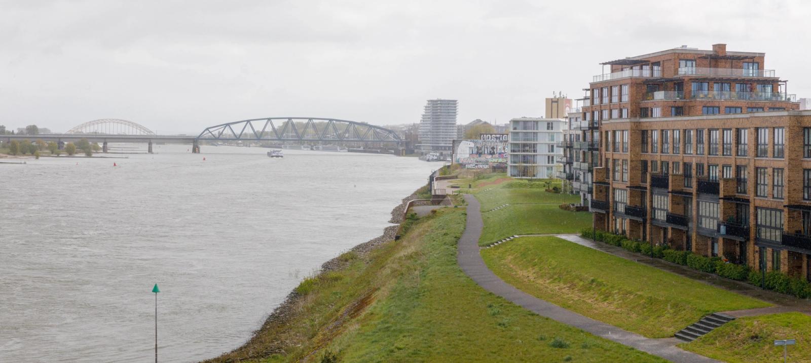 Links de rivier (de Waal), in het midden gras met een fietspad en rechts appartementgebouwen. 