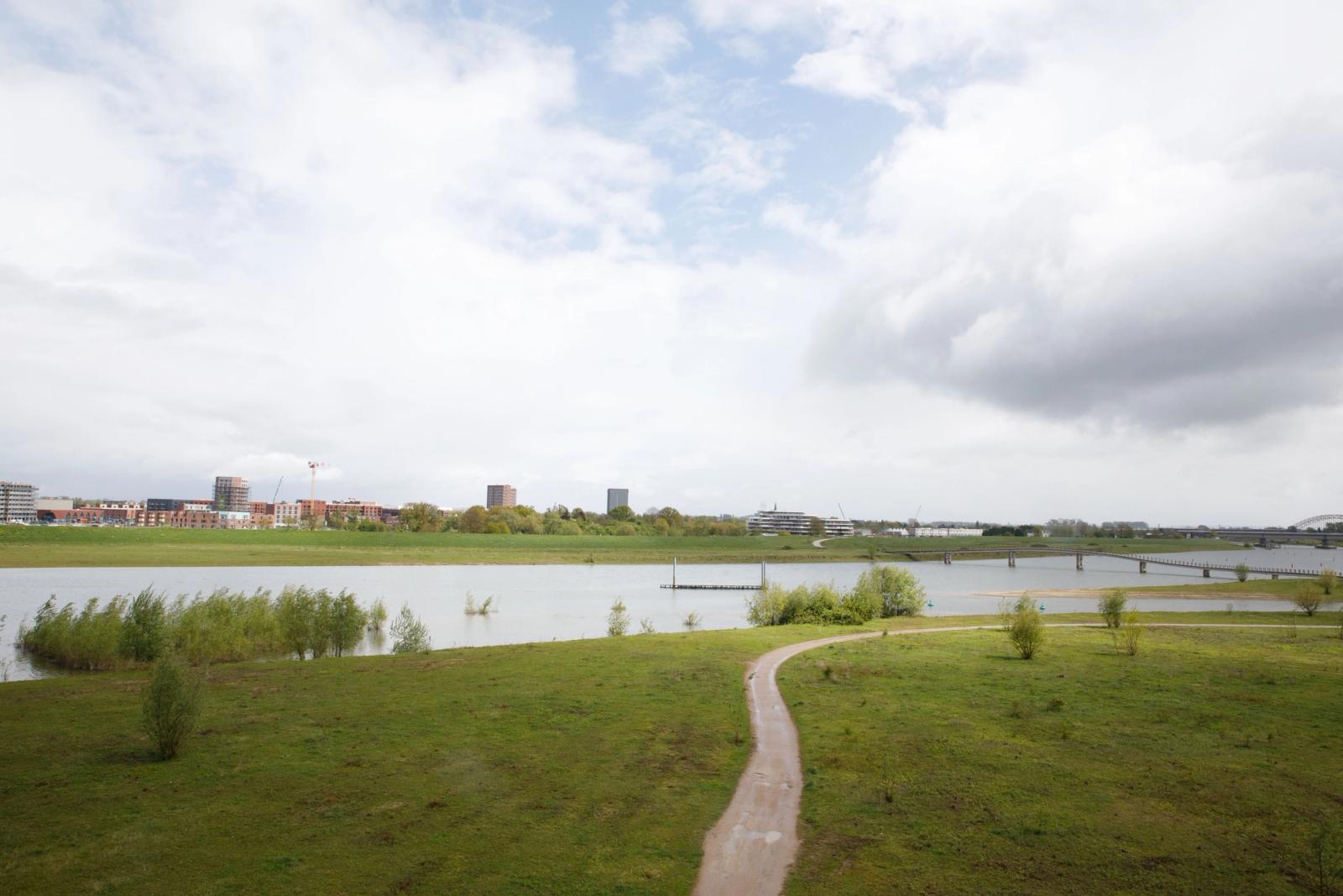 Een pad meandert door een grasveld heen aan de voorgrond. In het midden van het beeld is water (rivier) te zien. Op de achtergrond meer gras en bebouwing zoals flats, huizen en een hijskraan.