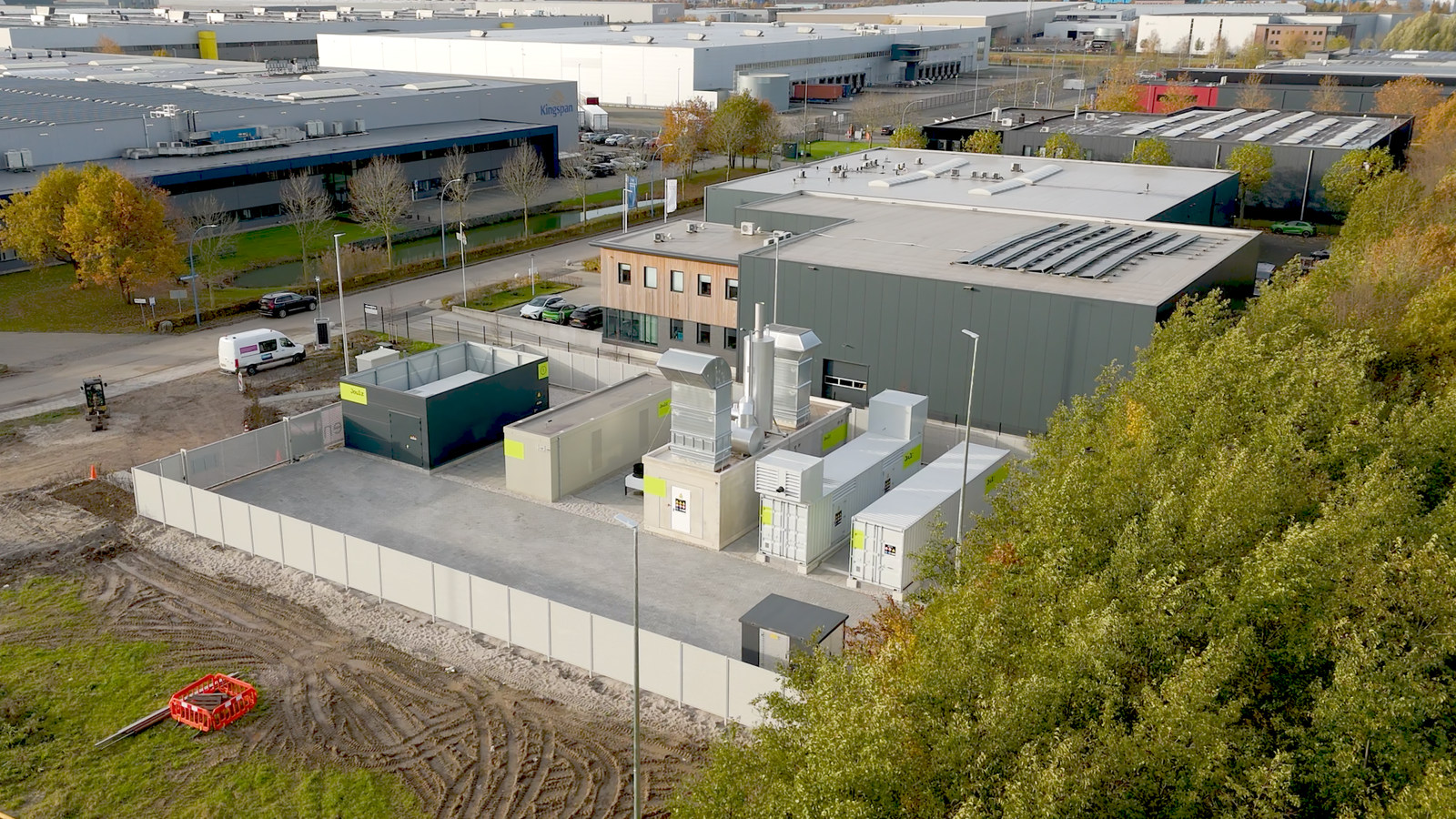 Rechts de toppen van bomen, linksonder zie je gras en modder/aarde. Daarachter zijn grijze gebouwen te zien die onderdeel maken van een bedrijventerrein. Aan de linkerkant van het bedrijventerrein is een autoweg te zien.