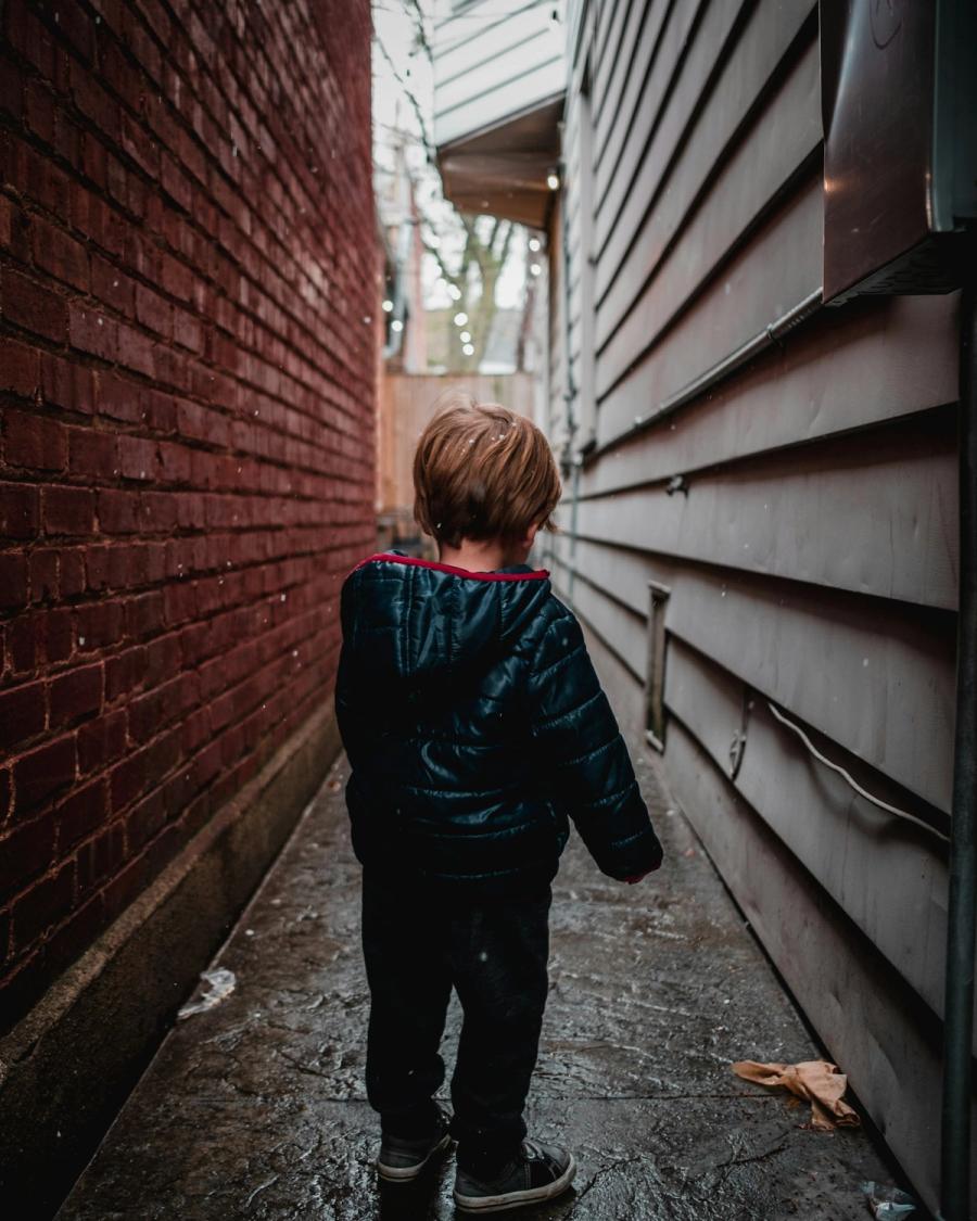 A toddler in an alley, turned partly away, capturing reflection and quiet resolve.