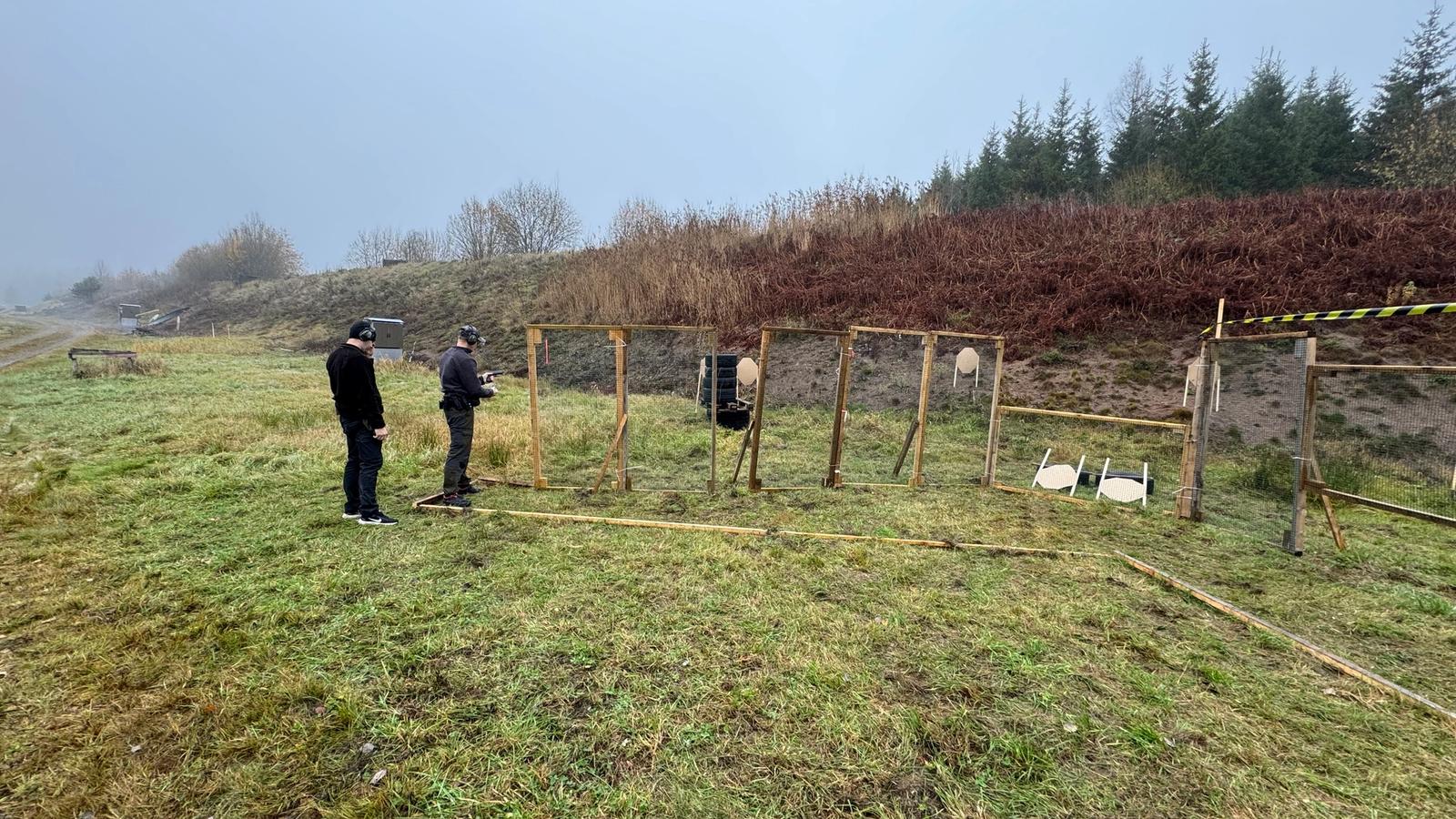 Skytt och skjutledare vid en IPSC-station utomhus, med papperstavlor och stålmål i träställningar på en dimmig skjutbana.