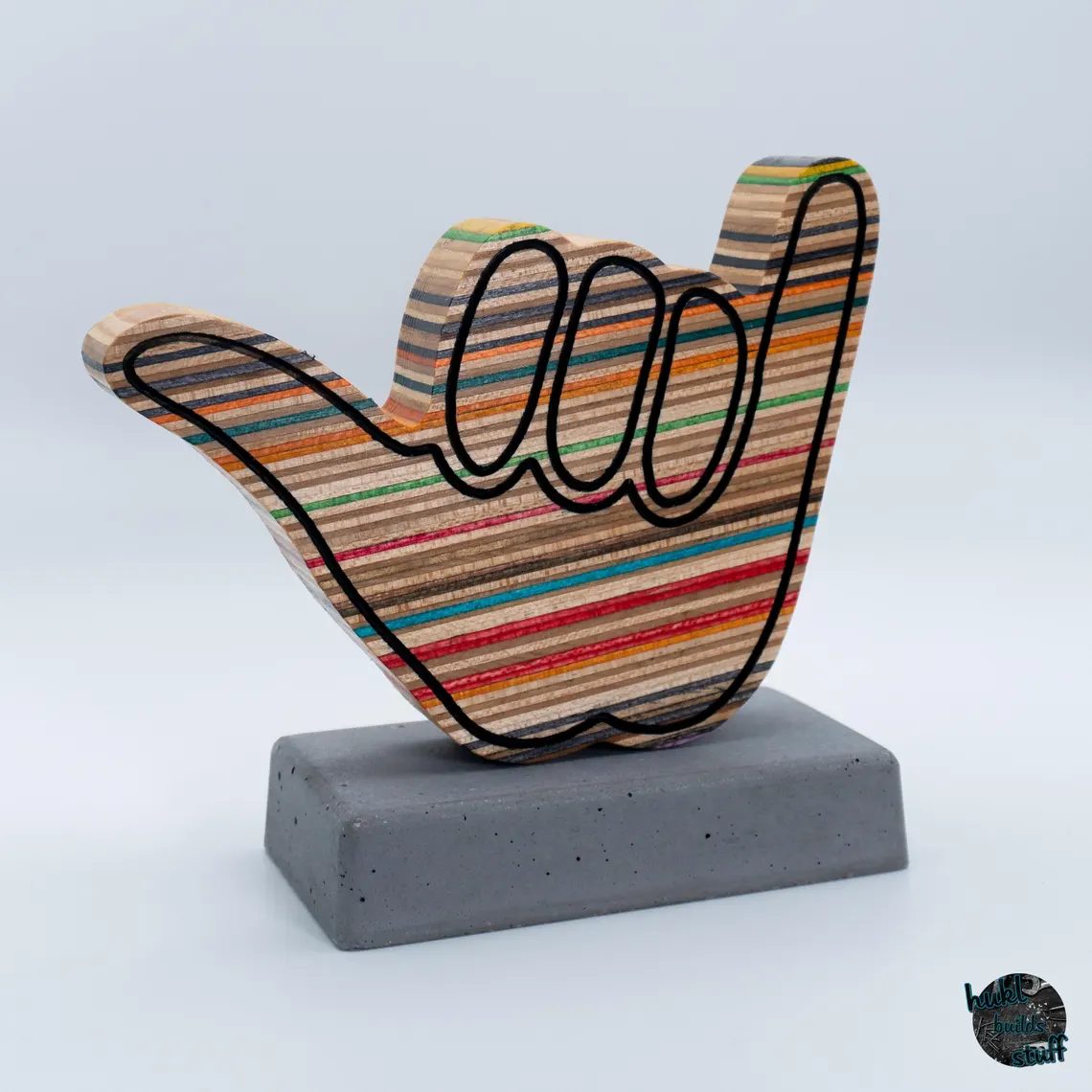A shaka hand sculpture made from recycled skateboard decks with engraved black details and a magnetic connection to a concrete base.