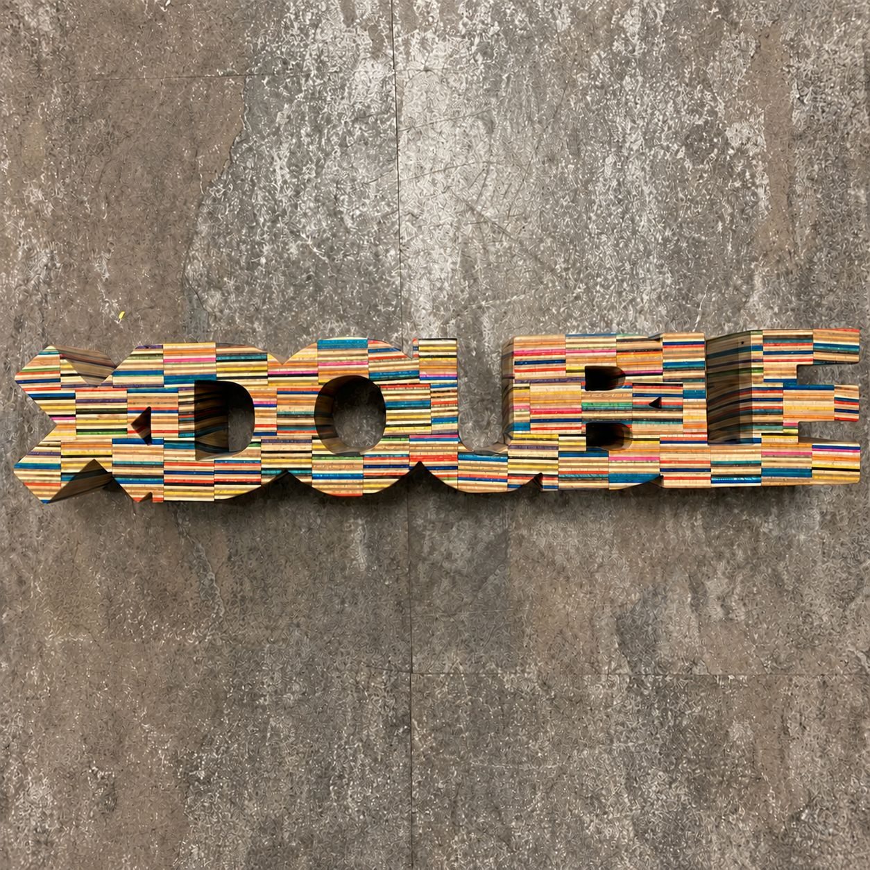 XDOUBLE lettering made from multicoloured recycled skateboards, photographed against a grey textured background.