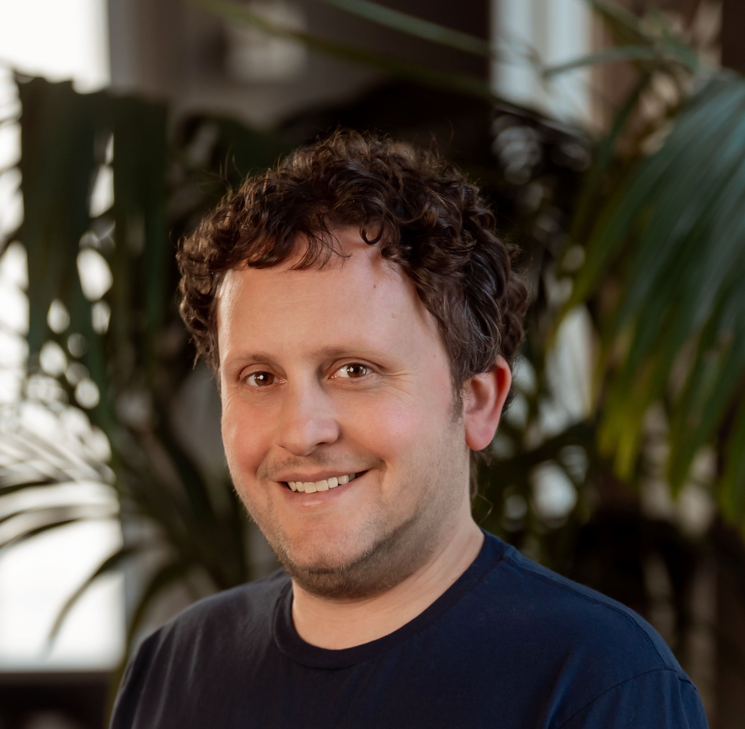 A smiling man with curly brown hair wearing a dark blue t-shirt.