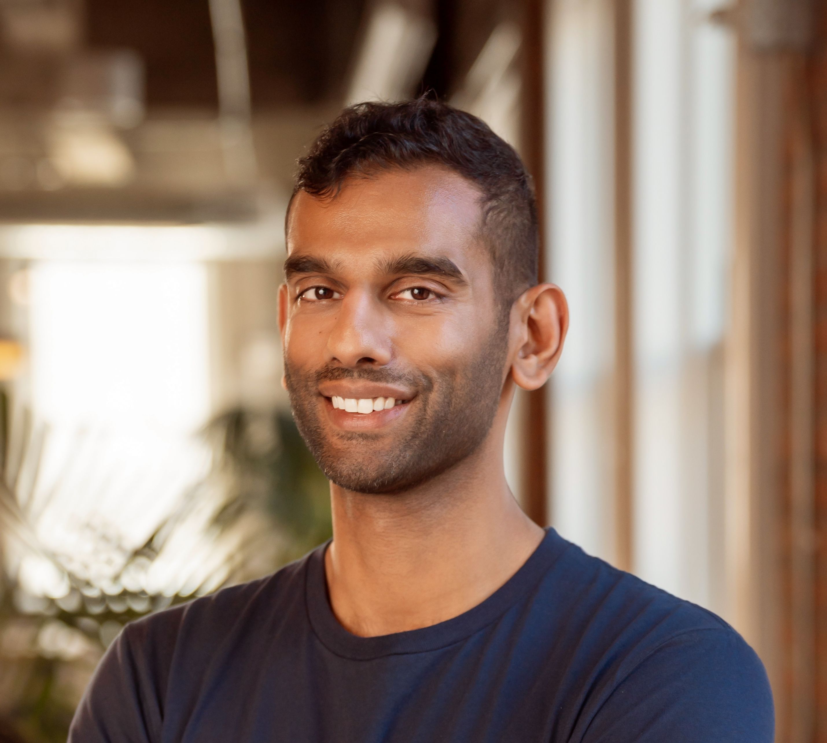 A smiling man with dark skin and dark hair wearing a navy t-shirt.