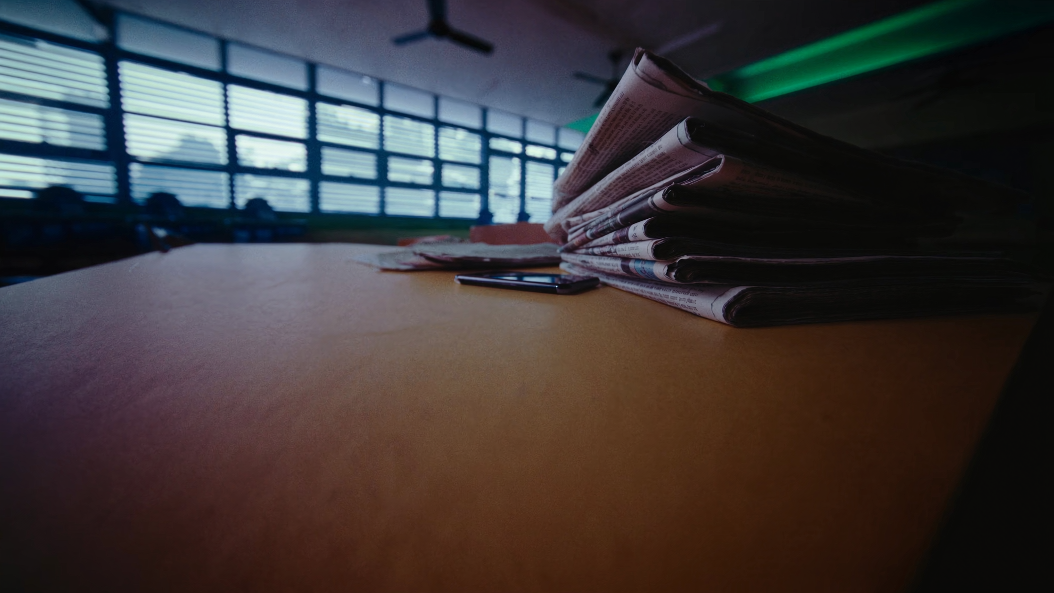 Timbunan surat khabar berlipat dan sebuah telefon pintar di atas meja kayu perpustakaan sekolah