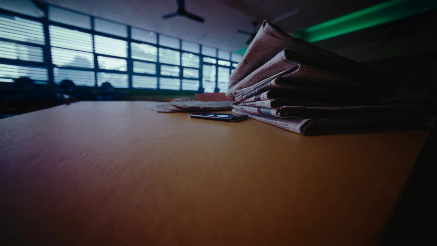 Timbunan surat khabar berlipat dan sebuah telefon pintar di atas meja kayu perpustakaan sekolah