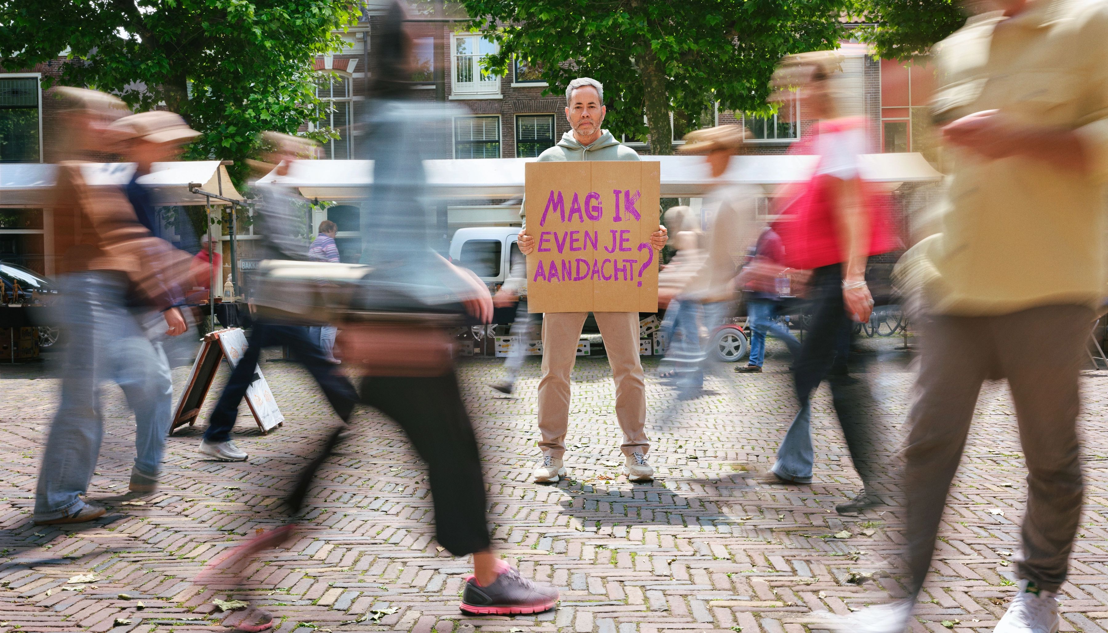 Een man, midden op een marktplein, heeft een bord in zijn hand met de tekst: Mag ik jouw aandacht?