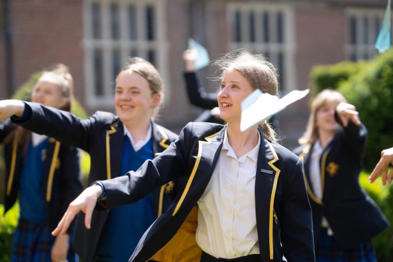 Students in school uniforms joyfully throwing paper planes outside a building.