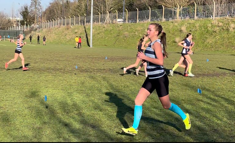A woman in athletic gear runs in a grassy outdoor setting during a race, with several other runners in the background.