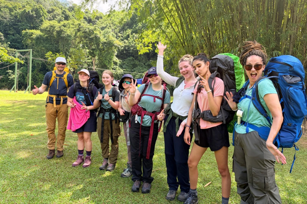 A group of World Challenge students and staff stand together with large backpacks, smiling and posing for the camera before setting off on their multi-day jungle trek through the Bornean rainforest. Surrounded by lush tropical greenery, the group look excited and ready for the adventure ahead.
