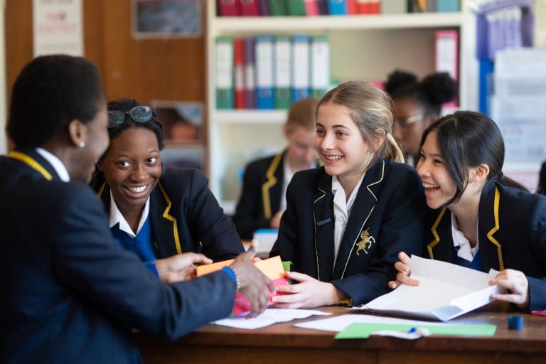 Four students in school uniforms sit at a table, smiling and chatting in a classroom.