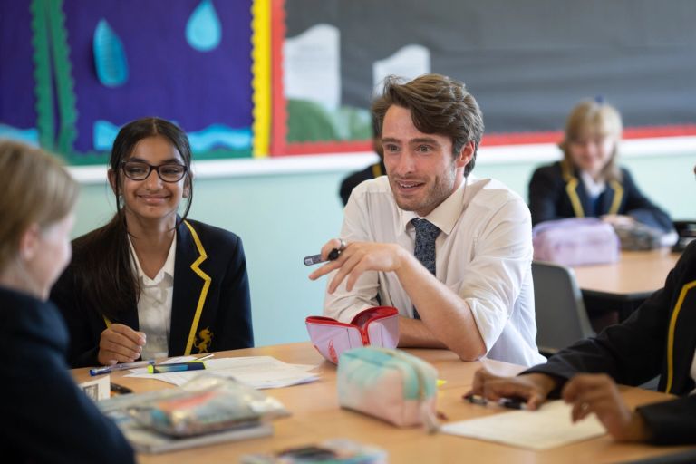 Teacher and students in school uniforms interact at a classroom table with papers and pencil cases.