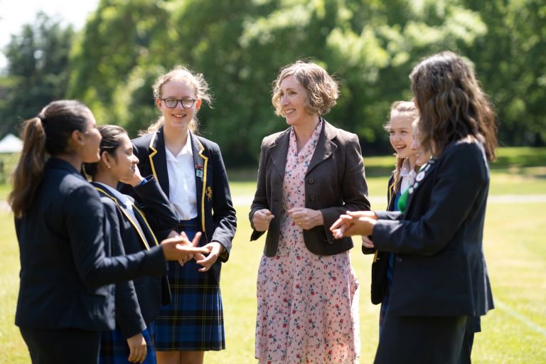 A group of students in uniforms standing and talking with a teacher outdoors on a sunny day.