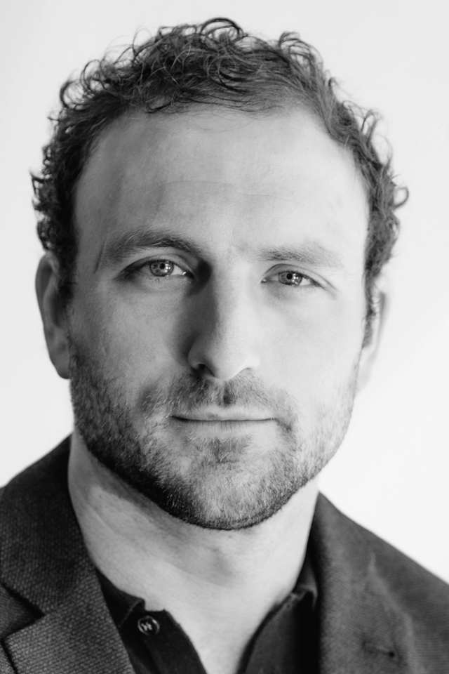 Portrait of Sam Allgood in a blue blazer against a white sunny background.