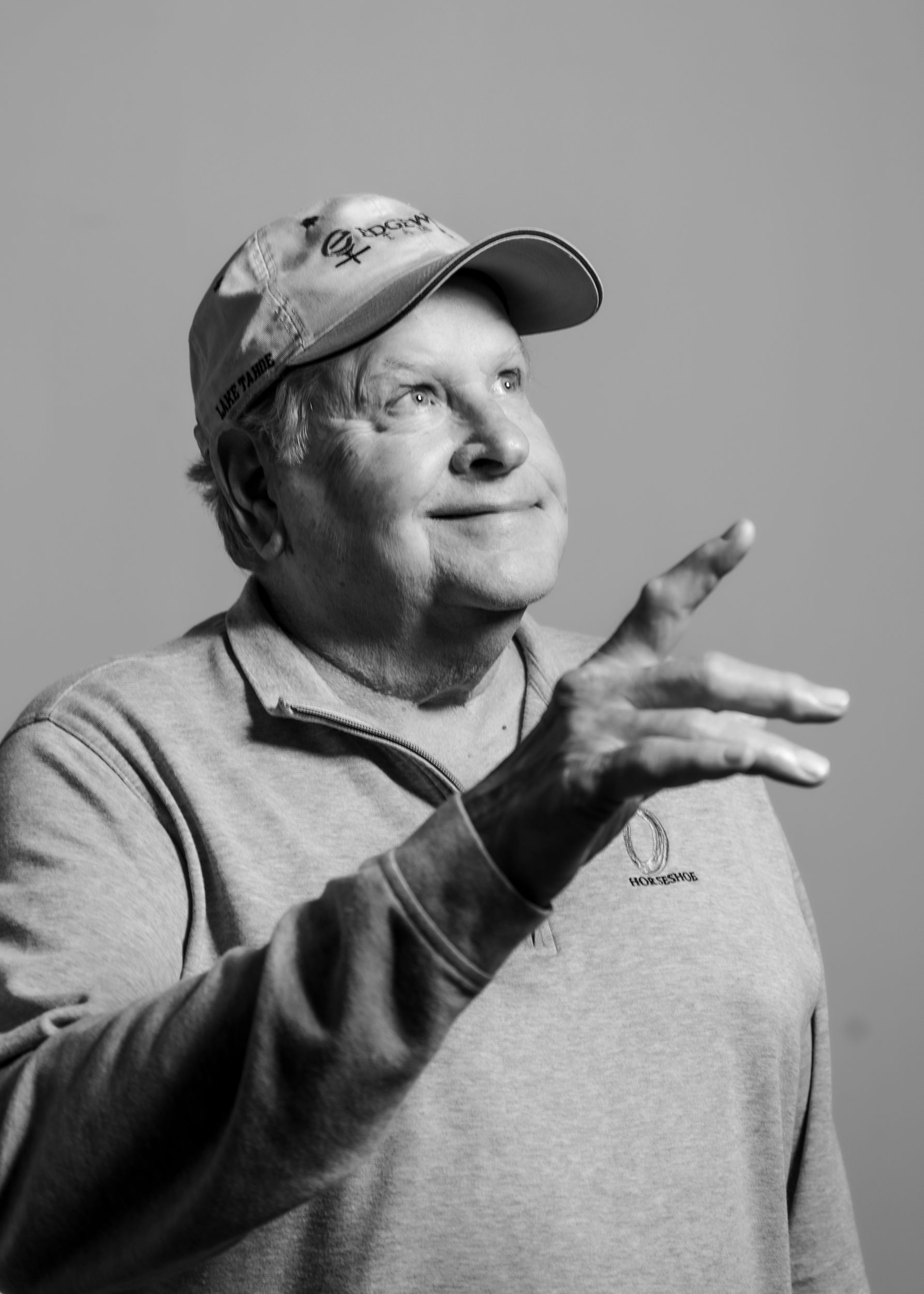 Black and white portrait of a grinning man, who is residential painter Gary Sanders, wearing a ball cap and pointing to the ceiling