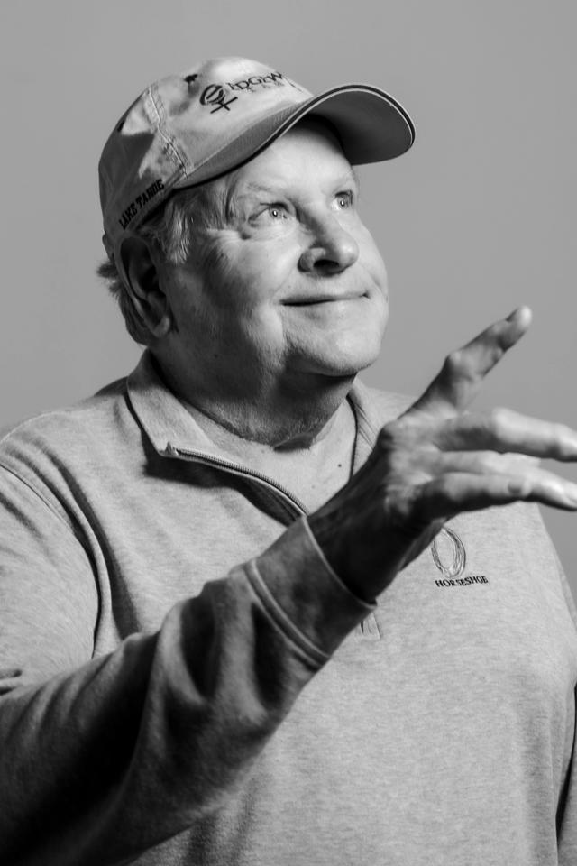 Black and white portrait of a grinning man, who is residential painter Gary Sanders, wearing a ball cap and pointing to the ceiling