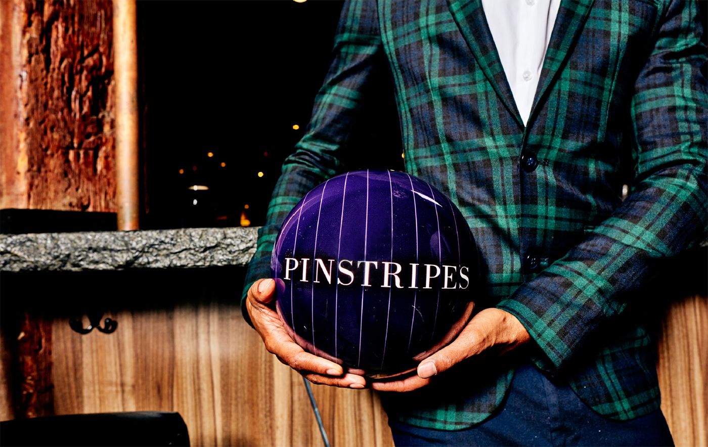 A guy in a quirky suit holding a bowling ball with the Pinstripes logo, smiling and ready to bowl, highlighting the fun, stylish side of the venue.