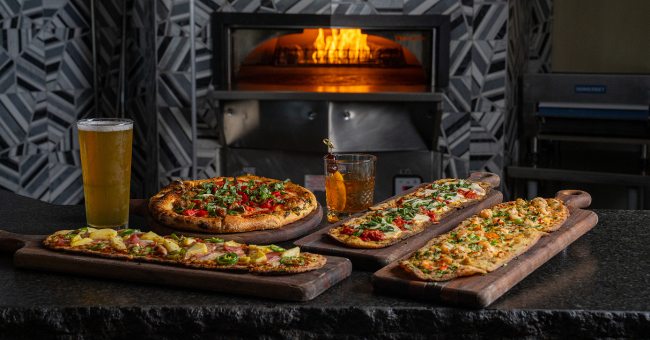Sausage and Peppers Pizza, Hawaiian, Margherita and Lobster flatbreads with beers in front of a wood-fired oven.