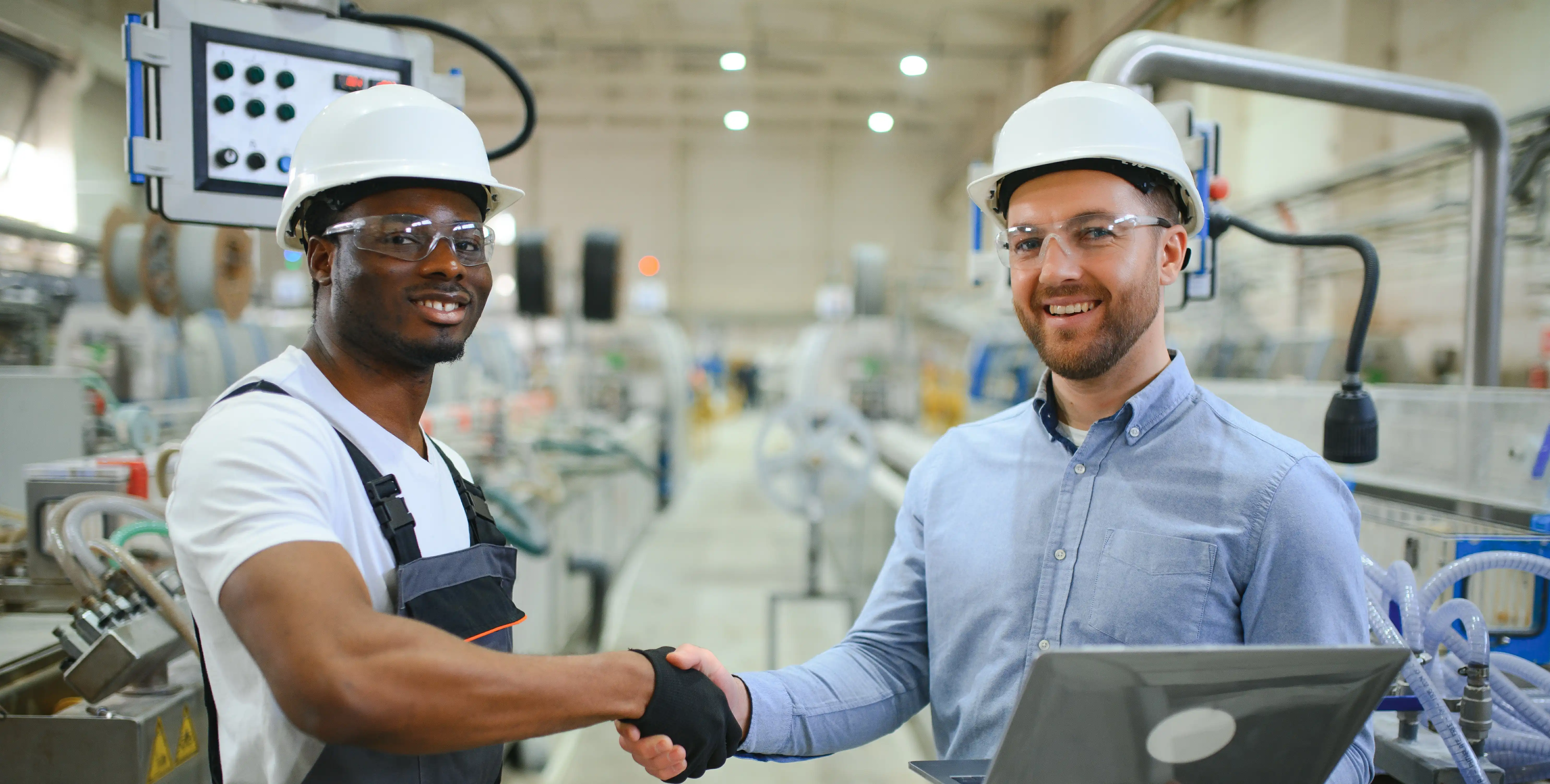 two factory workers with hard hats shaking hands and smiling at camera
