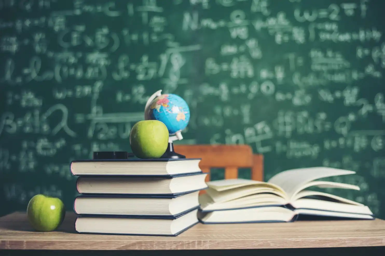 stack of books with green apples and globe and blackboard lesson in background